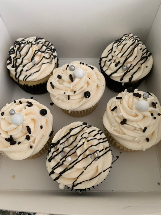 Six cupcakes with white frosting, black drizzle, and decorative elements in a box.